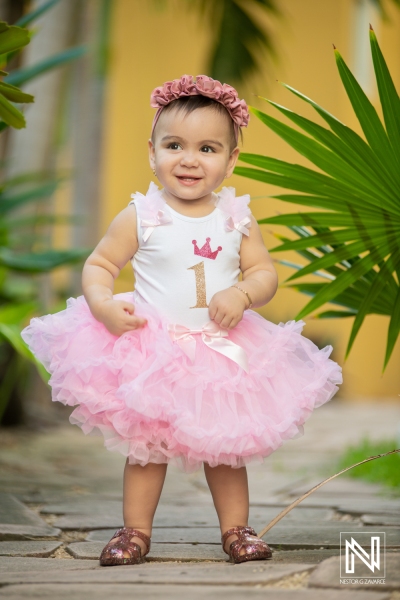 Cute toddler celebrating her birthday in Curacao, wearing a pink tutu and floral headband, surrounded by tropical plants and vibrant scenery