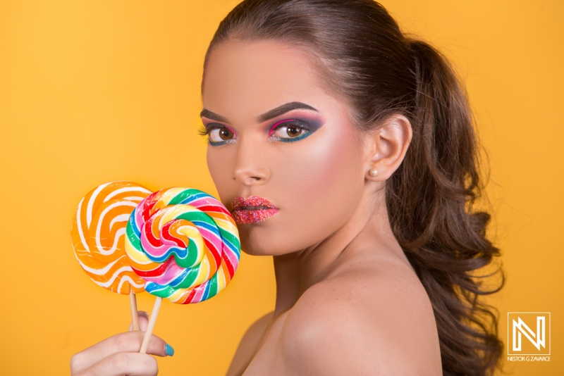 Model poses with colorful lollipops against bright yellow background in Curacao, showcasing vibrant makeup and playful style in a commercial setting