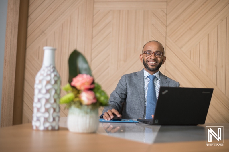 Professional man smiling at desk in Curacao office while working on laptop with decorative vase and flowers nearby