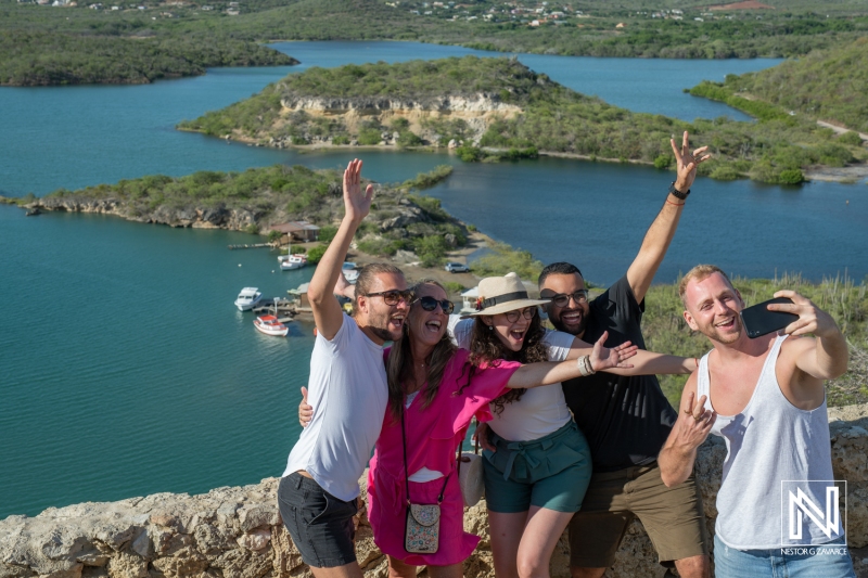 Group of friends enjoying a lively moment while taking a selfie at a scenic viewpoint in Curacao near a beautiful bay and lush landscape under bright sunlight