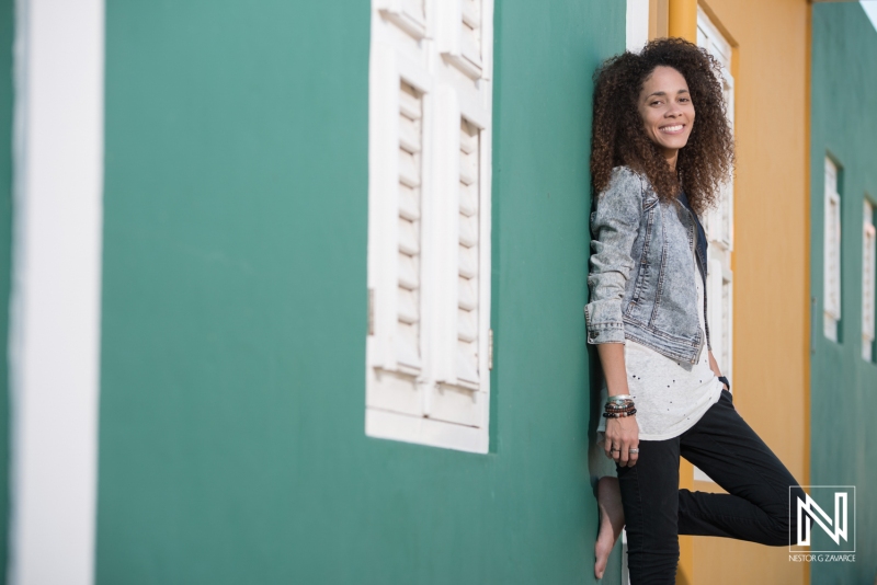 A Young Woman With Curly Hair Leans Against a Brightly Colored Wall in a Vibrant Urban Setting, Enjoying a Sunny Day While Showcasing Her Relaxed Style and Joyful Expression