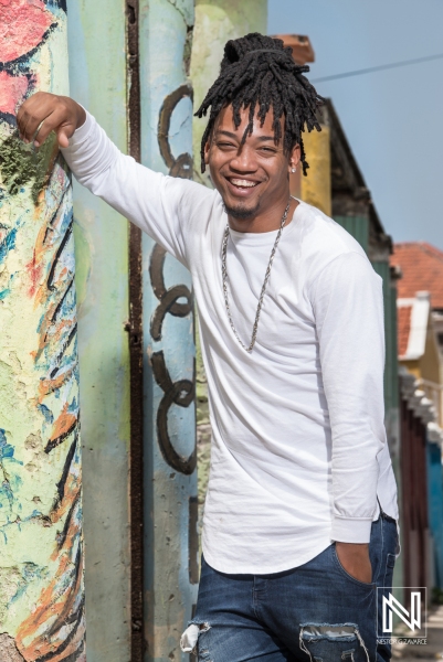 A Young Man With Dreadlocks Smiles While Leaning Against a Colorful Wall in a Vibrant Urban Area During Daylight Hours