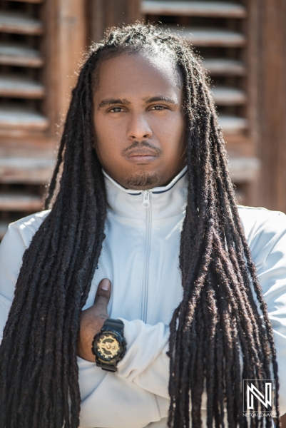 A Confident Man With Long Dreadlocks Poses Thoughtfully Outdoors in a Light Jacket Against a Rustic Wooden Background During the Afternoon