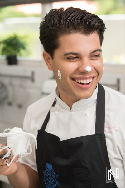 A Young Chef With a Playful Smile Holds a Bowl of Whipped Cream in a Modern Kitchen While Wearing a Black Apron During a Culinary Session