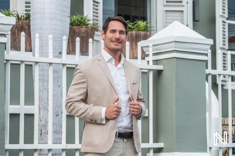 A Confident Man in a Beige Suit Stands Outside a Green House With a White Fence, Enjoying a Sunny Day While Adjusting His Blazer in a Suburban Neighborhood