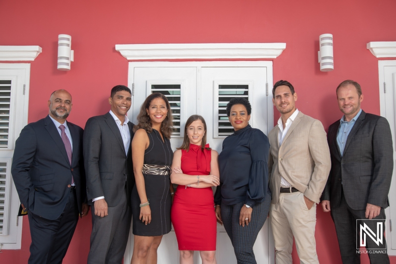 A Diverse Group of Professionals Gathers in Formal Attire in Front of a Bright Red Wall With White Shutters, Engaging in Collaboration at a Networking Event in a Warm, Sunny Location