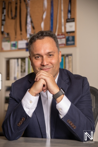 A Professional Man With a Friendly Smile Poses Confidently at an Office Desk Surrounded by Various Awards and Accomplishments in a Contemporary Workspace