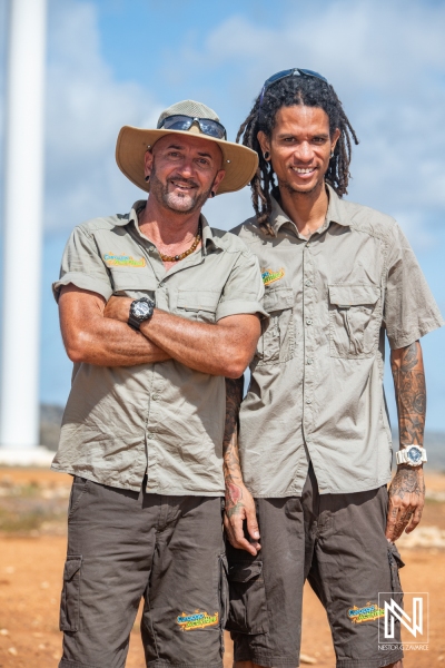 Two Men Dressed in Safari Attire Pose Confidently Outdoors Near a Wind Turbine on a Sunny Day in a Rural Area, Showcasing a Sense of Camaraderie and Adventure