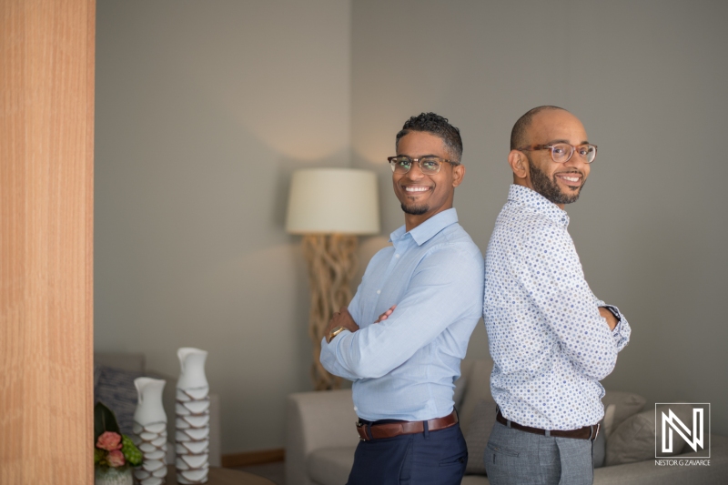 Two Men Pose Confidently Back to Back in a Stylish Modern Living Room, Showcasing Their Friendship and Professional Attire in a Relaxed Indoor Setting