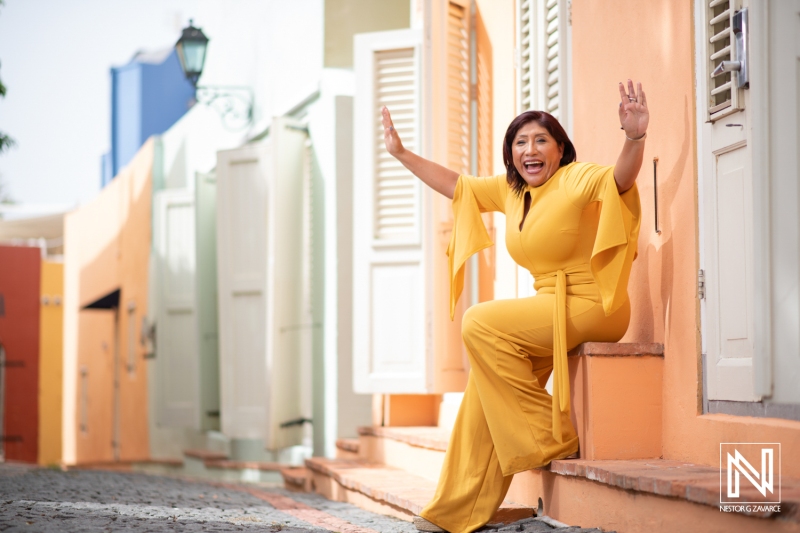 A Woman Joyfully Poses in a Bright Yellow Outfit, Showcasing Vibrant Urban Architecture While Enjoying a Sunny Day on a Colorful Street