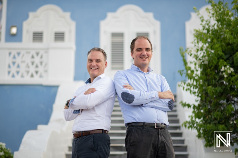 Two Smiling Men Stand Confidently on Outdoor Stairs in Front of a Light Blue Building During Daytime, Dressed in Casual Business Attire, Exuding a Sense of Camaraderie and Professionalism