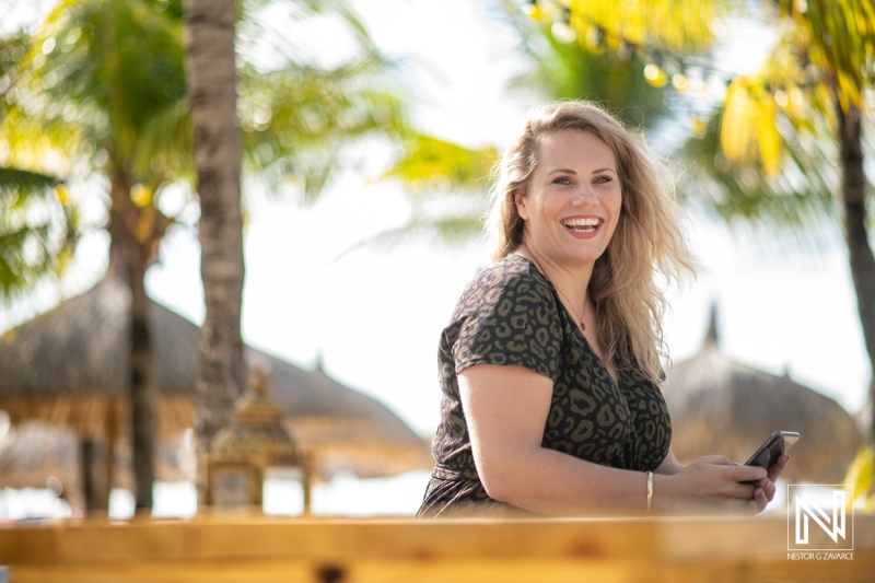 A Woman Smiles While Sitting by the Beach Under Palm Trees During a Warm Sunny Day, Enjoying Her Time at a Tropical Resort