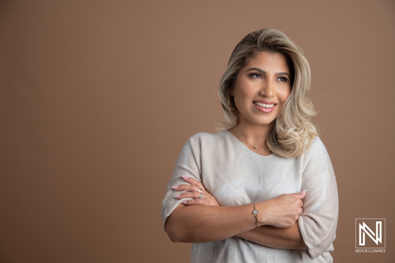 A Smiling Young Woman With Blonde Hair Crosses Her Arms While Standing Against a Neutral Brown Background in a Well-Lit Indoor Setting