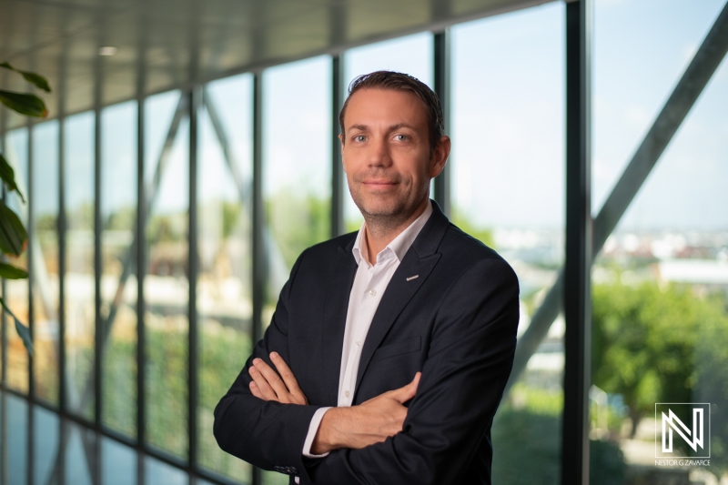 Professional Headshot of a Man in a Smart Blazer, Posing Confidently Indoors With Large Windows Showcasing Greenery and a Bright Sky, Conveying a Sense of Professionalism and Approachability