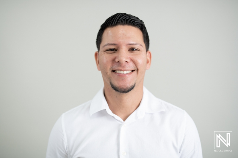 A Smiling Man Dressed in a White Shirt Poses Against a Plain Background in a Modern Indoor Setting, Showcasing a Friendly Demeanor and Engaging Personality in Natural Light