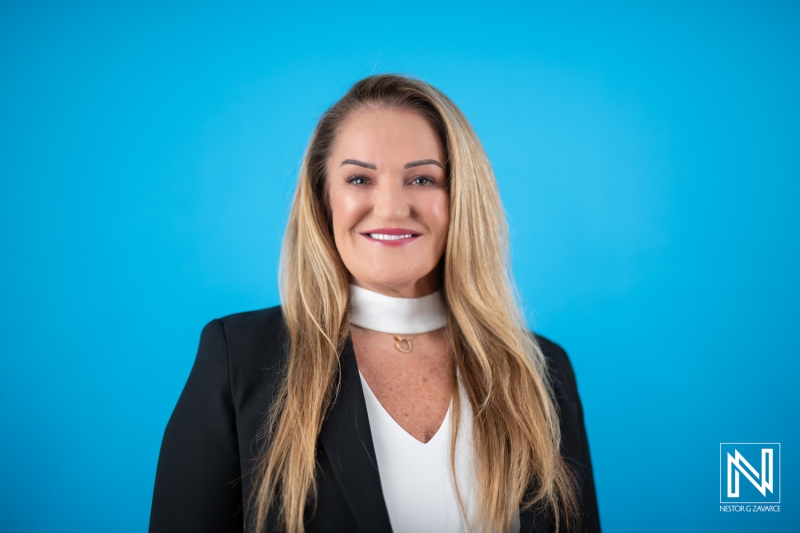 A Professional Woman in a Black Blazer Smiles Confidently Against a Bright Blue Background, Showcasing Her Approachable Demeanor in an Office Setting