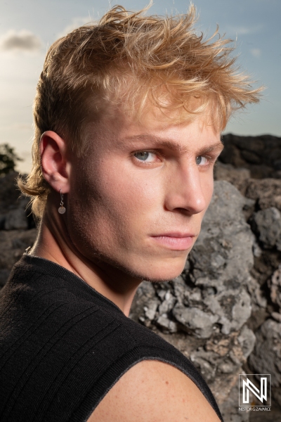 A Young Man With Short Blond Hair Poses Confidently Against a Rocky Background During Sunset, Showcasing a Striking Gaze and Casual Attire