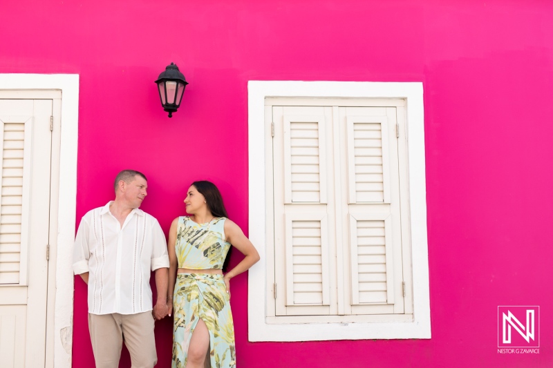 Couple Holding Hands in Front of a Vibrant Pink Wall With White Shutters While Enjoying a Sunny Day in a Charming Neighborhood