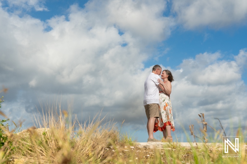 A Couple Shares a Romantic Moment on a Sunny Day Near the Coastline, Surrounded by Fluffy Clouds and Tall Grass, Capturing Their Love in a Picturesque Outdoor Setting