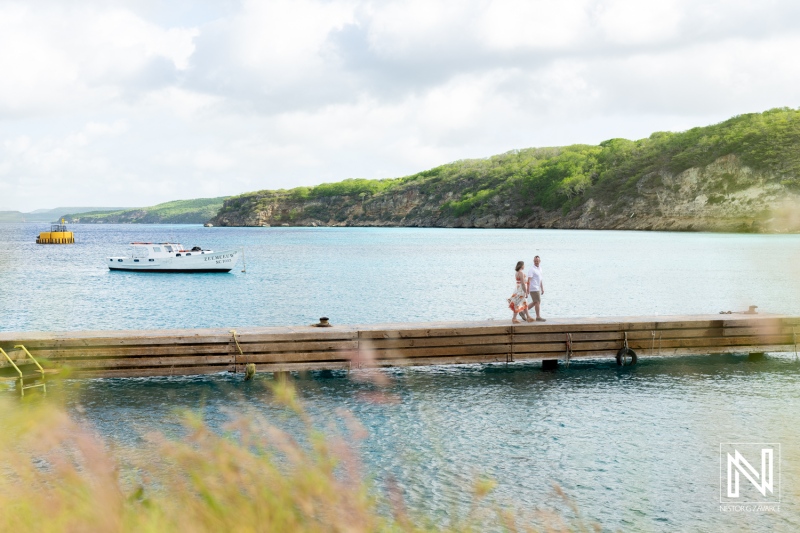 Couple Strolling Along the Wooden Pier With a Boat Anchored in a Calm Bay, Surrounded by Lush Hills Under a Partly Cloudy Sky