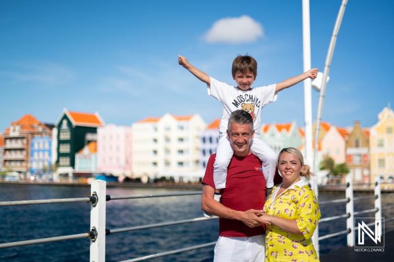 Family enjoys a sunny day in Curacao with colorful buildings in the background while the child playfully sits on the parent's shoulders