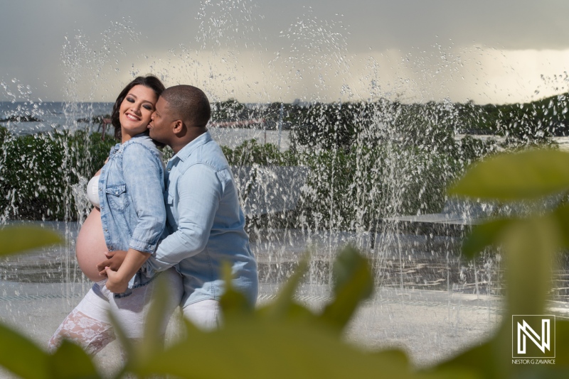 Expecting parents share a joyful moment by a fountain in Curacao during a sunny day in their maternity photoshoot