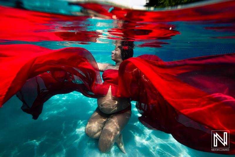 Maternity shoot in Curacao featuring a pregnant woman underwater with flowing red fabric in a tranquil pool setting