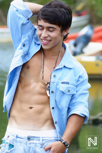 Young man posing confidently by the water wearing a light blue jacket and displaying a toned physique during a sunny day outdoors