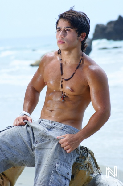 A young man poses confidently on a rock by the beach with the ocean waves in the background, enjoying a sunny day and showing off his toned physique