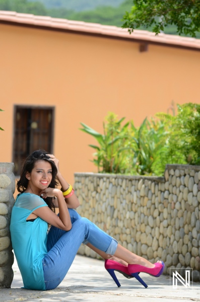 Young woman in casual attire poses confidently by a stone wall in a sunny outdoor environment, enjoying a relaxed moment surrounded by greenery