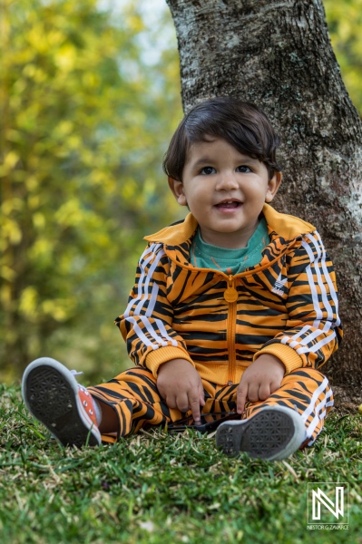 Young child in tiger-striped tracksuit sitting under a tree in a sunny park, enjoying a playful moment outdoors during the day