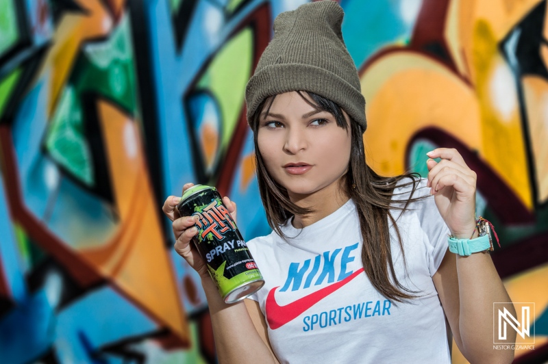 A young woman poses confidently in an urban setting wearing a casual outfit with vibrant graffiti art in the background while holding a spray can in the early afternoon