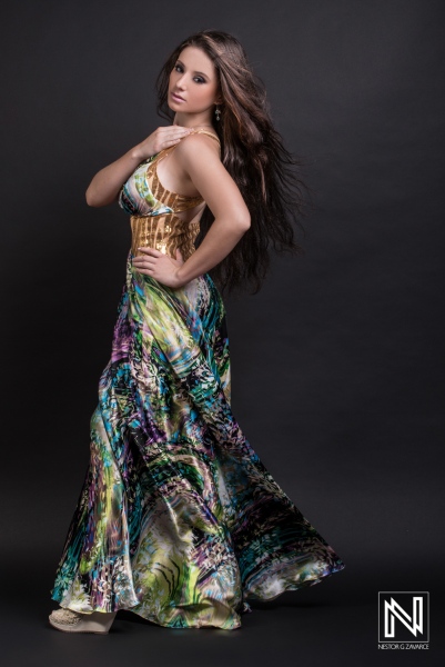 A young woman poses confidently in a colorful gown with a flowing design, showcasing elegance and grace against a dark backdrop in a studio setting