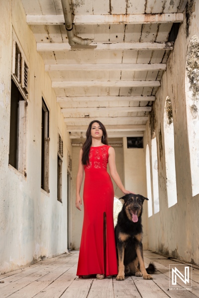 Young woman in a red dress stands beside a large dog in a rustic corridor with wooden floors and weathered walls in the afternoon light