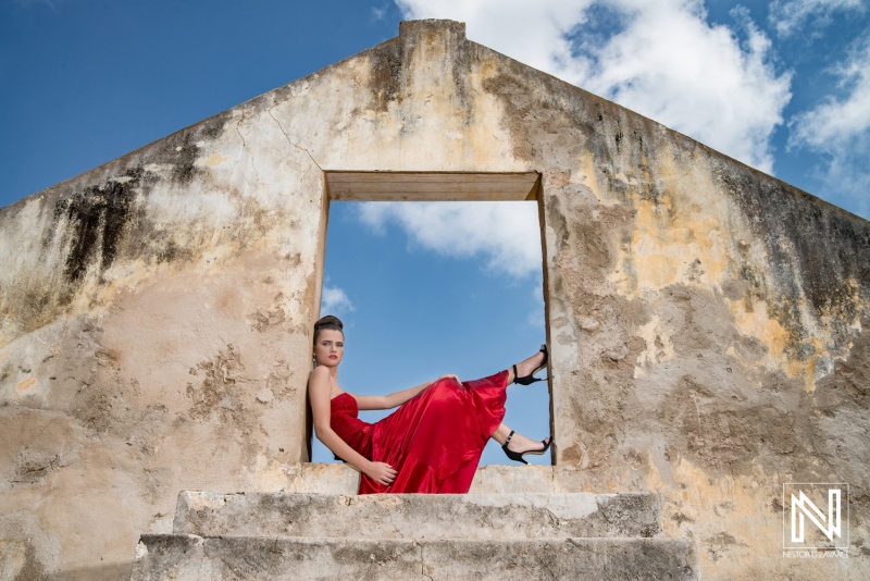 A woman in a flowing red dress poses elegantly at an abandoned building under a bright blue sky with scattered clouds during daylight