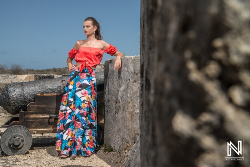 A model poses in vibrant clothing against a historic backdrop featuring old cannons near a scenic coastal area under a clear blue sky