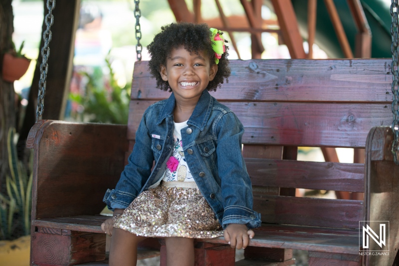 A happy girl with curly hair wearing a denim jacket and sparkly skirt sitting on a swing in a colorful playground during a sunny day