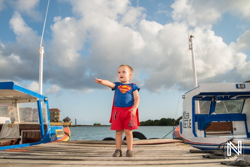 A young child dressed as a superhero stands confidently on a pier, pointing towards the sky while two colorful boats rest nearby on a sunny day