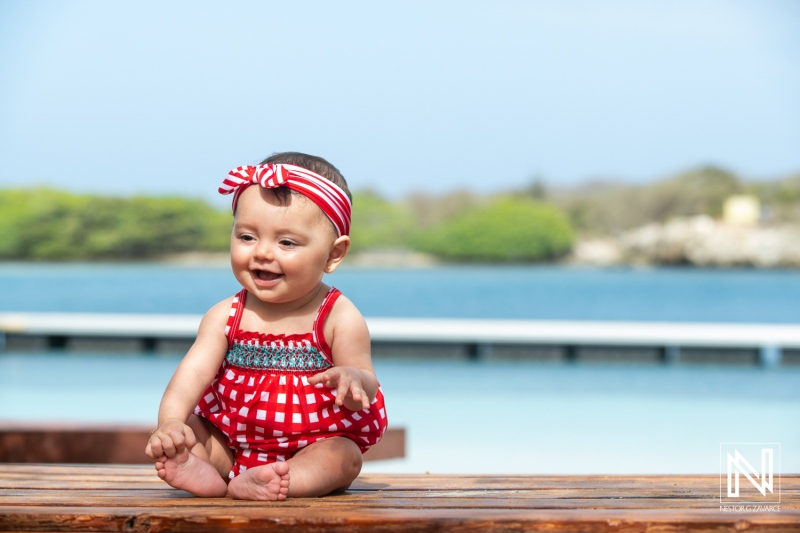 A joyful baby girl in a red and white checkered swimsuit sits by the water, enjoying a sunny day at the beach with a bright smile on her face