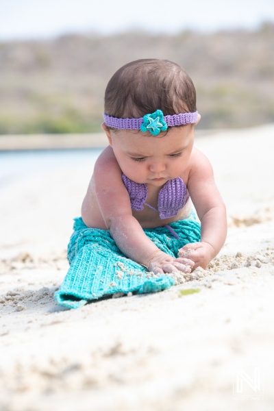 A baby girl exploring the sandy beach while dressed in a colorful mermaid costume on a sunny day by the water