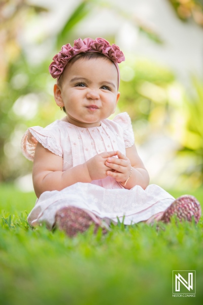 A young girl sitting on green grass in a park, wearing a pink dress and flower headband, making a playful expression during a sunny afternoon