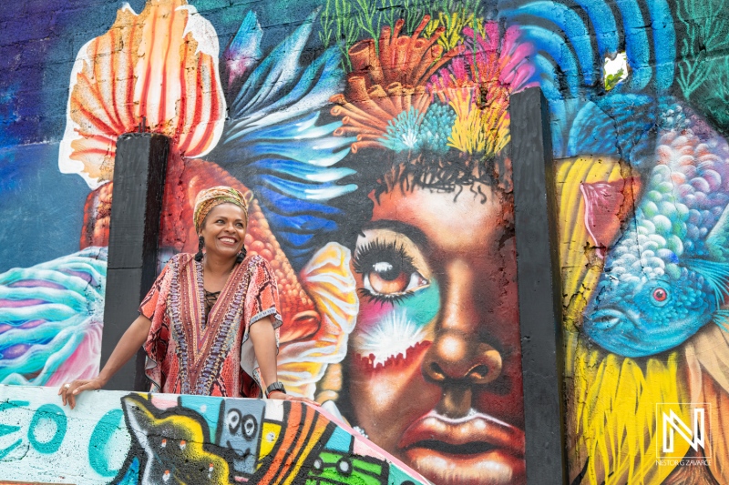 A woman smiles joyfully in front of vibrant street art featuring colorful fish and a striking portrait in a lively urban setting