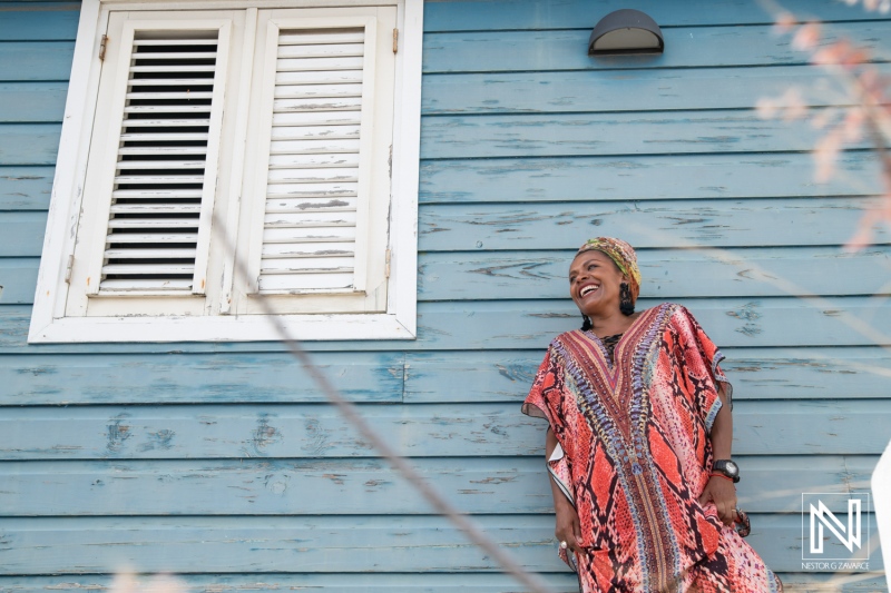 A joyful woman in a colorful dress stands in front of a blue wooden house with shuttered windows under bright sunlight in a vibrant neighborhood