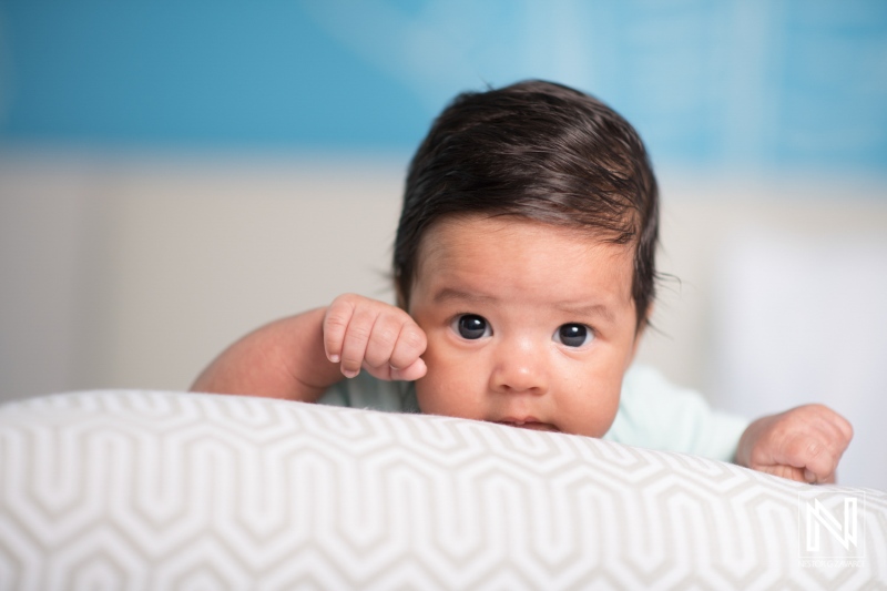 A baby with dark hair is playfully peeking over a patterned cushion, displaying curiosity and playfulness in a bright indoor setting