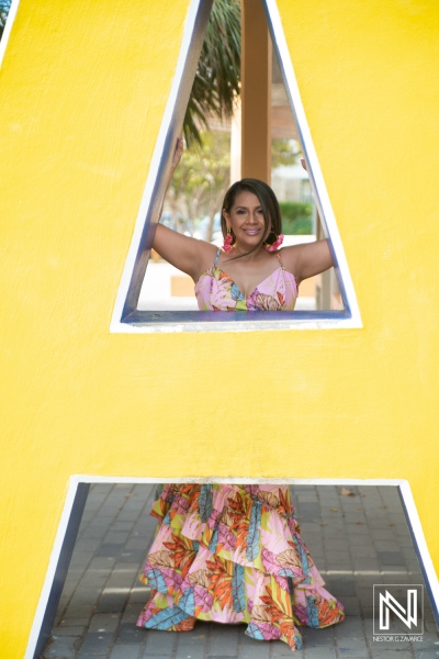 A woman in a colorful dress poses joyfully through a large yellow letter A at a vibrant location during the daylight