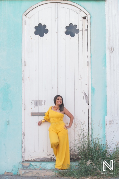 Young woman in a bright yellow outfit poses confidently by a white wooden door with floral designs on a pastel blue wall during golden hour