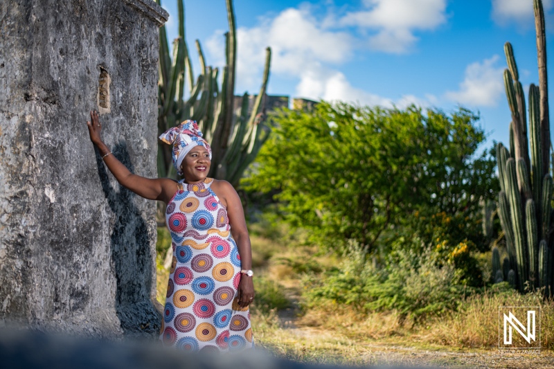 A woman dressed in a colorful traditional outfit stands against an old stone wall surrounded by cactus plants during a bright sunny day in a rural setting