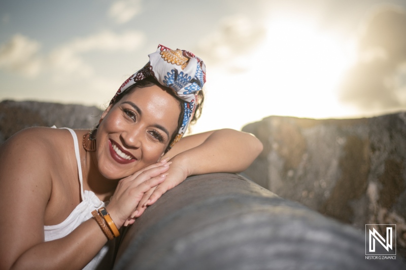 A cheerful woman with a colorful head wrap enjoys the seaside at sunset, resting on a stone structure while smiling brightly and soaking in the warm sunlight