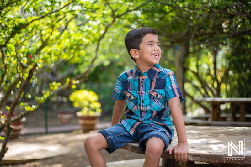 A young boy enjoying a sunny day outdoors while sitting on a wooden table in a garden surrounded by lush greenery and colorful plants in the afternoon