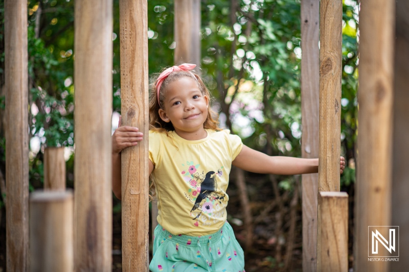 A young girl joyfully playing among wooden posts in a sunny outdoor setting, surrounded by greenery during a bright afternoon